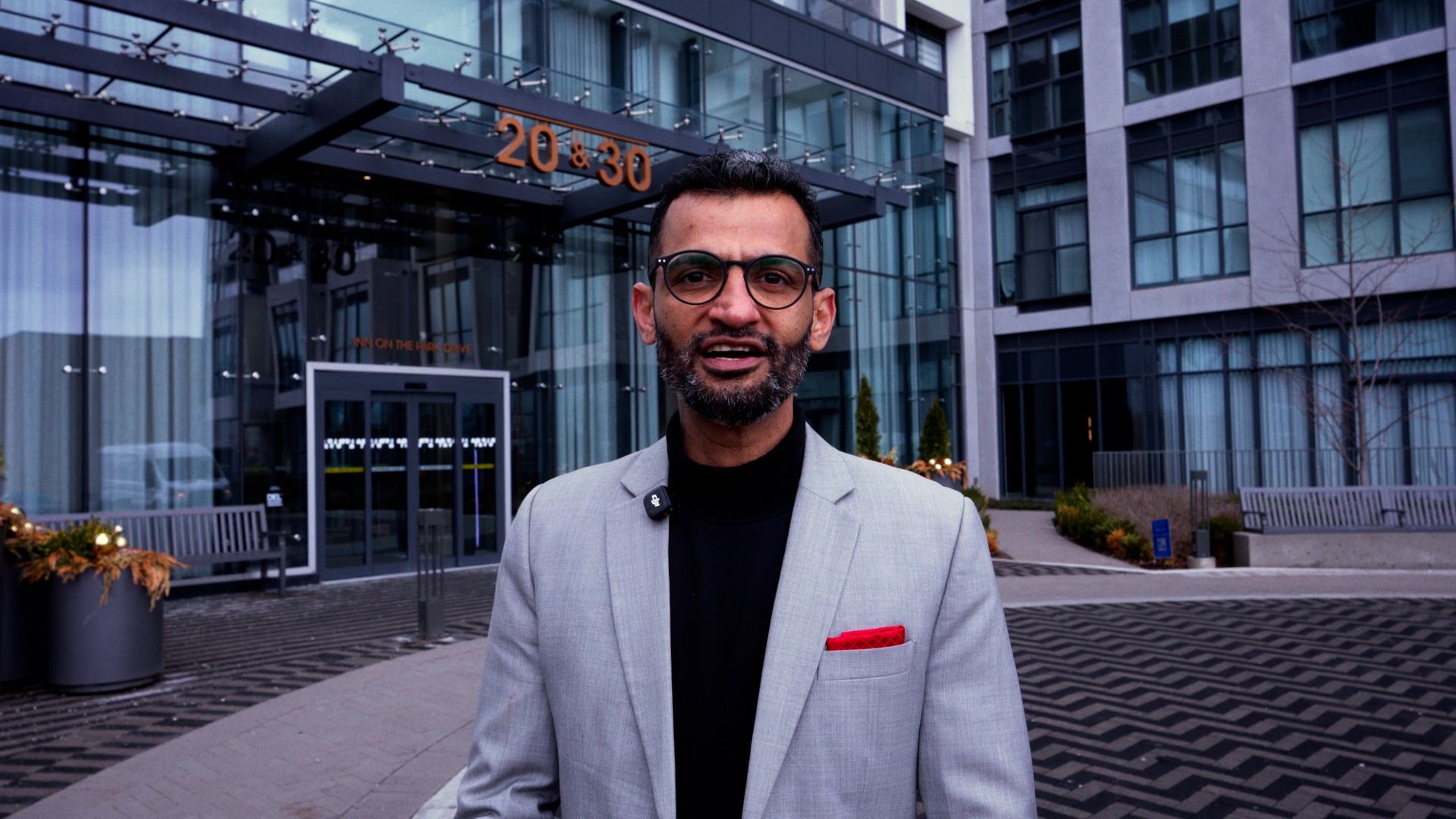 A man wearing glasses and a grey jacket outside a modern building.